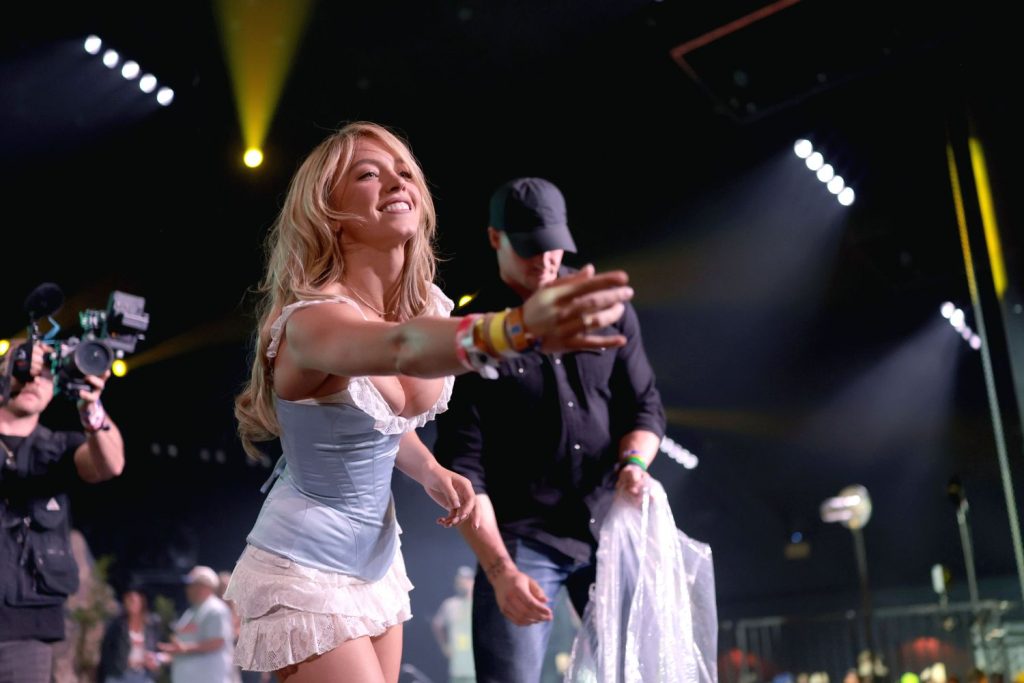 Sydney Sweeney in a light blue corset and white skirt reaches toward the audience on stage, smiling, while several people and cameras surround her under bright stage lights.