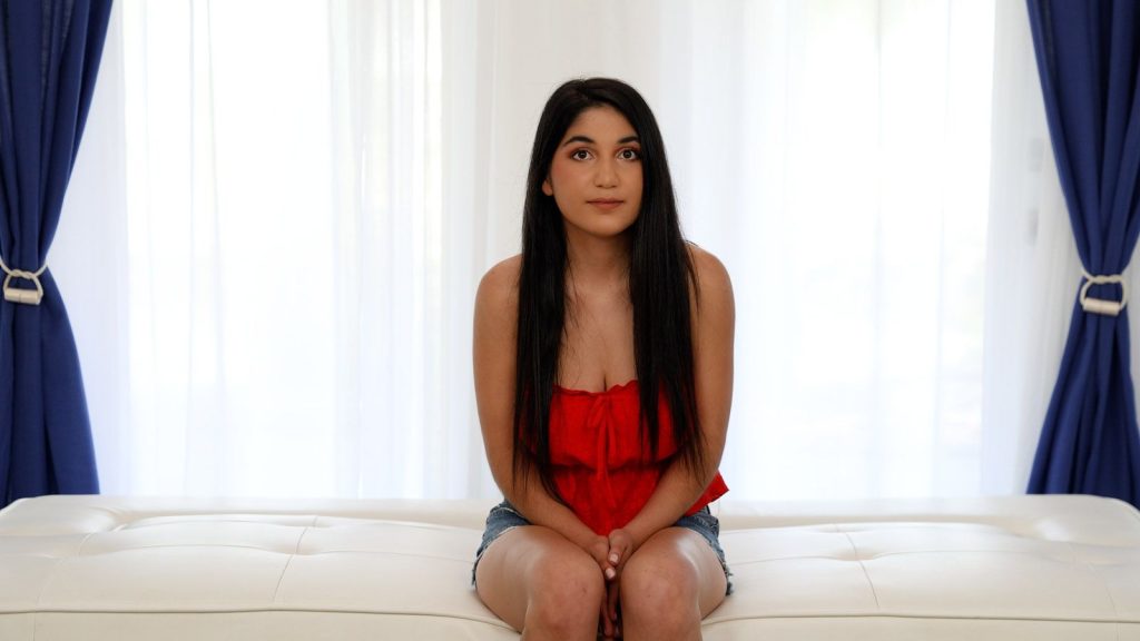 A young woman with long dark hair, wearing a red sleeveless top and denim shorts, sits on a white bench in a bright room with blue curtains and white sheer drapes behind her. She looks into the camera.