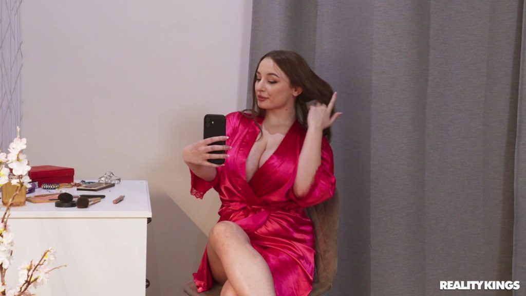 A woman wearing a bright pink satin robe sits on a chair, holding a smartphone up for a selfie while touching her hair. She is next to a white vanity with makeup items. Curtains and a wall are in the background.
