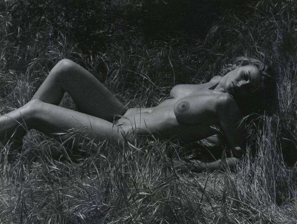 A nude woman lying on her side in tall grass, chest raised and resting on her elbows. She turns her head to the side. The image is in black and white.