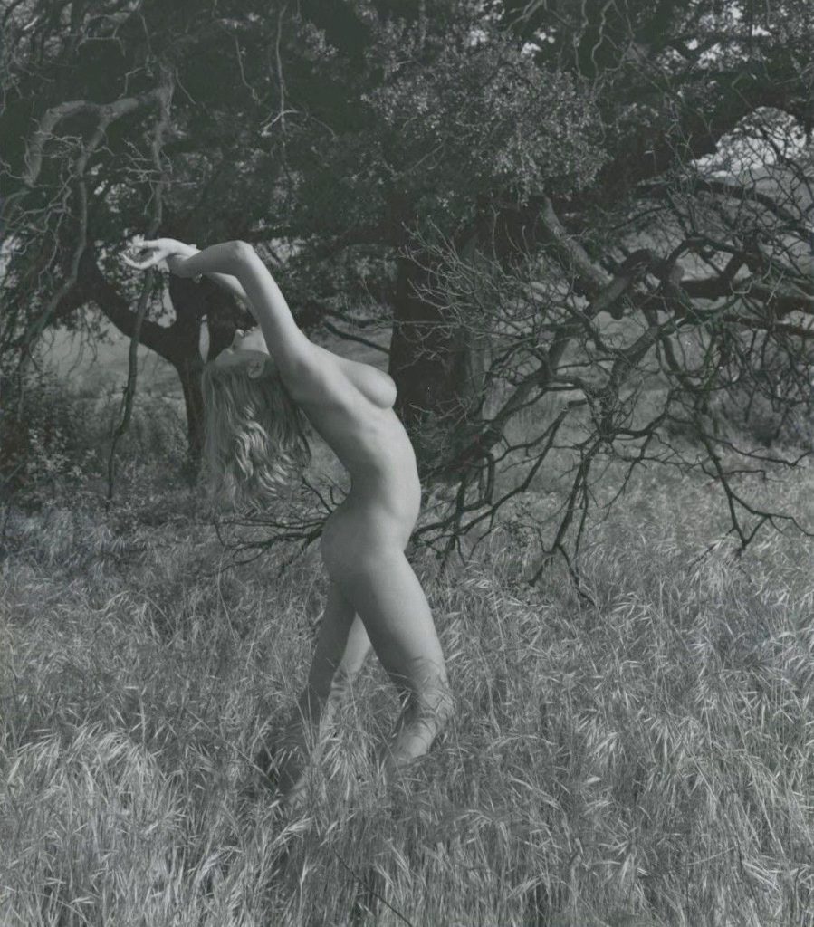 A nude woman stands in tall grass, arching their back and stretching their arms overhead. Leafy trees with twisted branches fill the background, creating a natural setting. The image is in black and white.