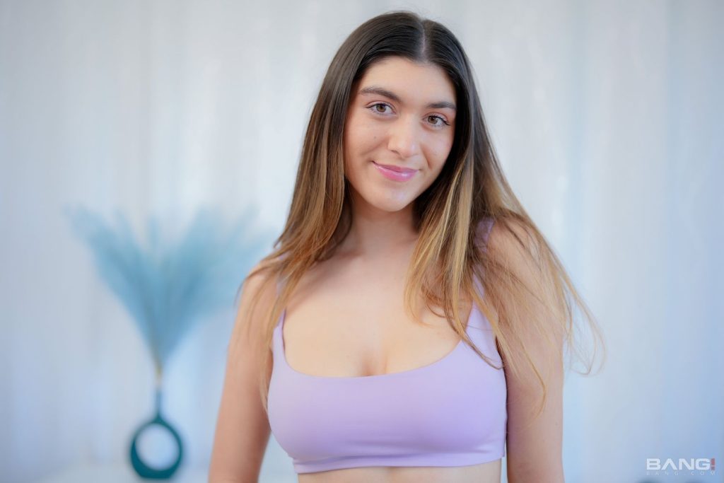 A woman with long brown hair wearing a light purple tank top smiles softly at the camera, standing indoors with a blurred blue vase and light background behind her.