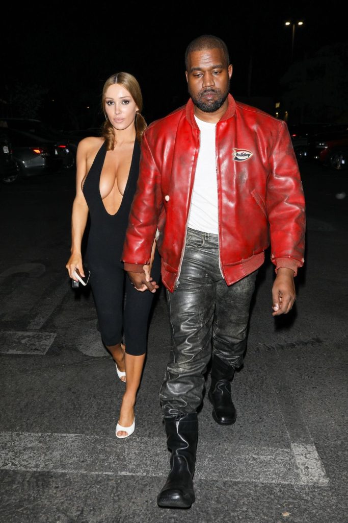 A woman in a black jumpsuit and white heels walks holding hands with a man wearing a red leather jacket, gray pants, and black boots at night in a parking lot #7.