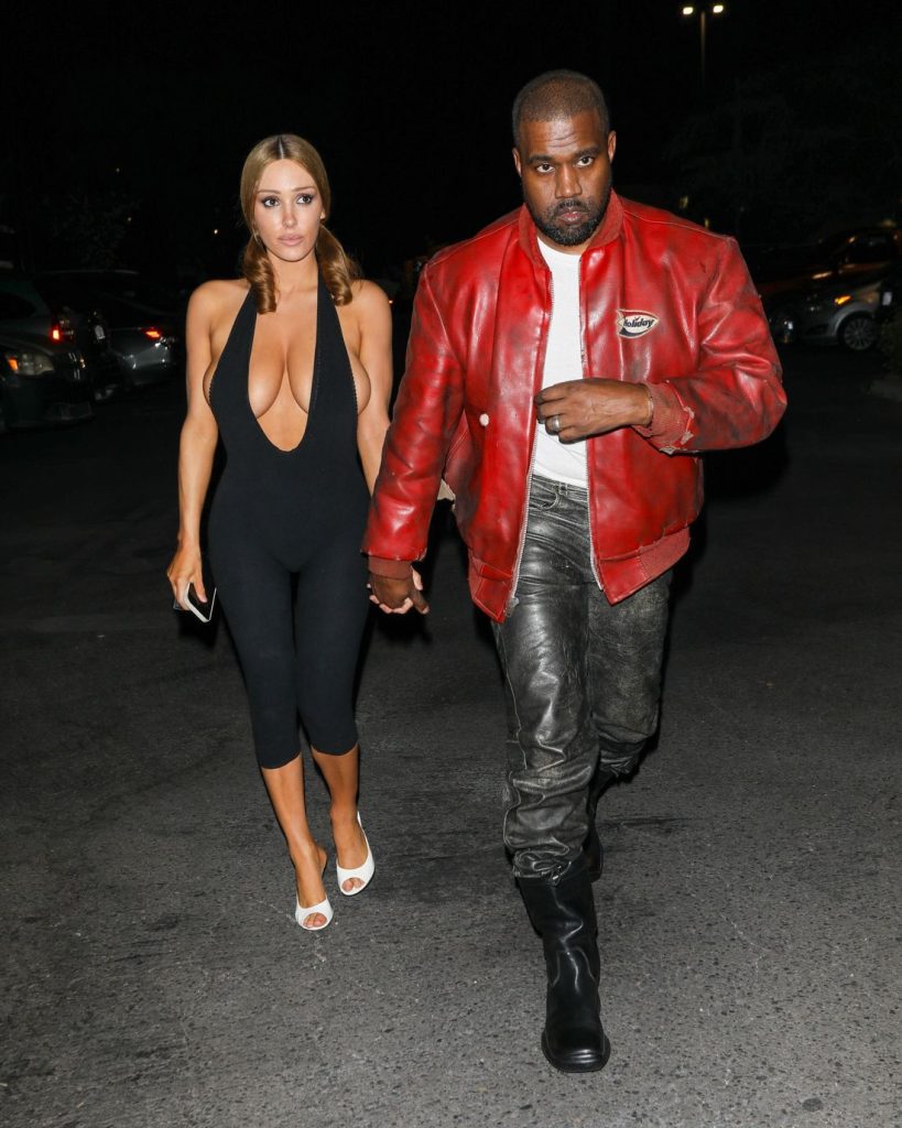 A woman in a black jumpsuit and white heels walks holding hands with a man wearing a red leather jacket, gray pants, and black boots at night in a parking lot #9.