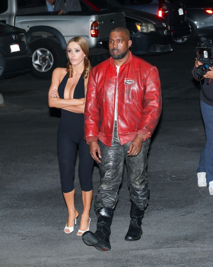 A man in a red leather jacket and black pants walks next to a woman wearing a black jumpsuit and white heels. They are outside at night in a parking lot, with cars and a cameraman in the background.