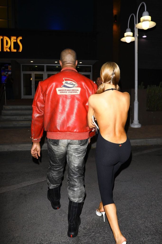 A man in a red leather jacket and jeans walks beside a woman wearing black pants and a backless top, holding his arm as they walk at night near a building with bright lights. View is from behind the couple.