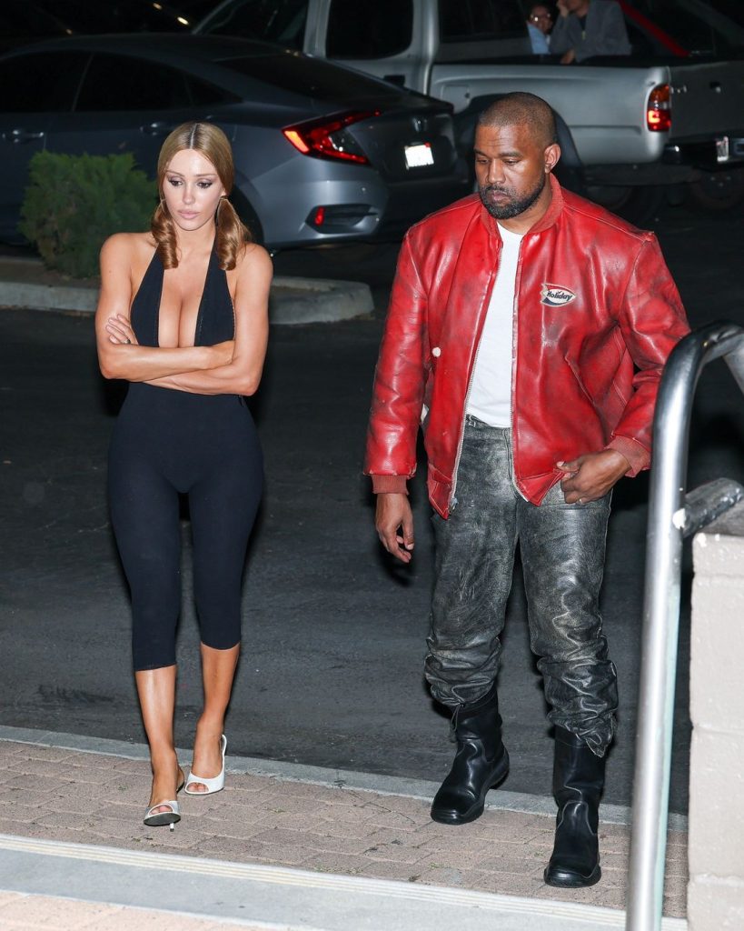 A woman in a black sleeveless jumpsuit and white heels walks with her arms crossed beside a man wearing a red leather jacket, black shirt, metallic pants, and black boots in a parking lot at night.