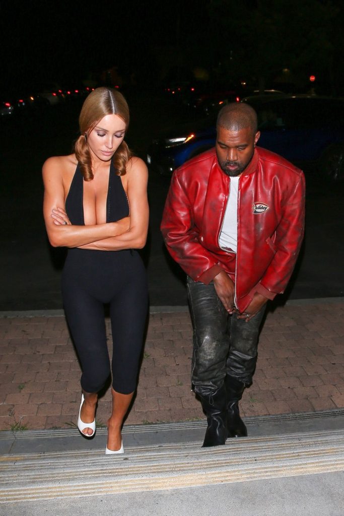 A woman in a black sleeveless jumpsuit and white heels walks with her arms crossed beside a man wearing a red leather jacket, black shirt, metallic pants, and black boots in a parking lot at night #2.