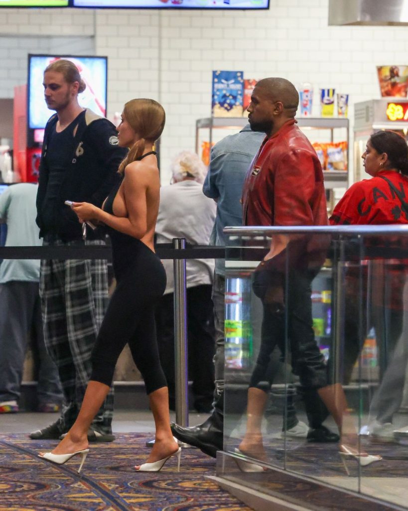 Three people stand in line at a movie theater concession stand. The woman in front wears a black outfit and white heels, while the two men behind her wear casual clothes. Popcorn and snacks are visible in the background.