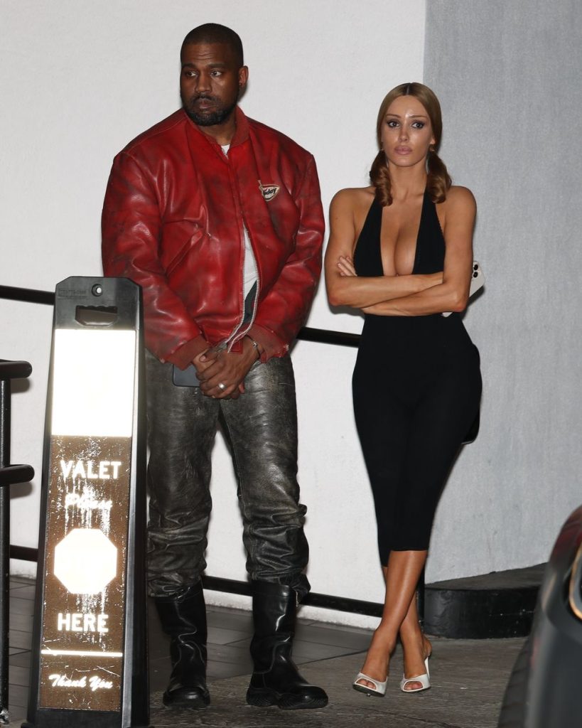 A man in a red jacket and black pants stands next to a woman in a sleeveless black dress and white heels. The woman has her arms crossed. They are near a valet parking sign outside at night.