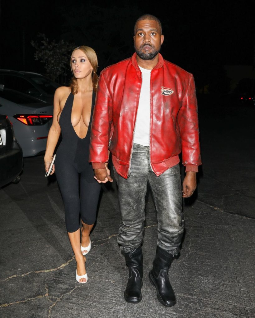 A woman in a black jumpsuit and white heels walks holding hands with a man wearing a red leather jacket, gray pants, and black boots at night in a parking lot.