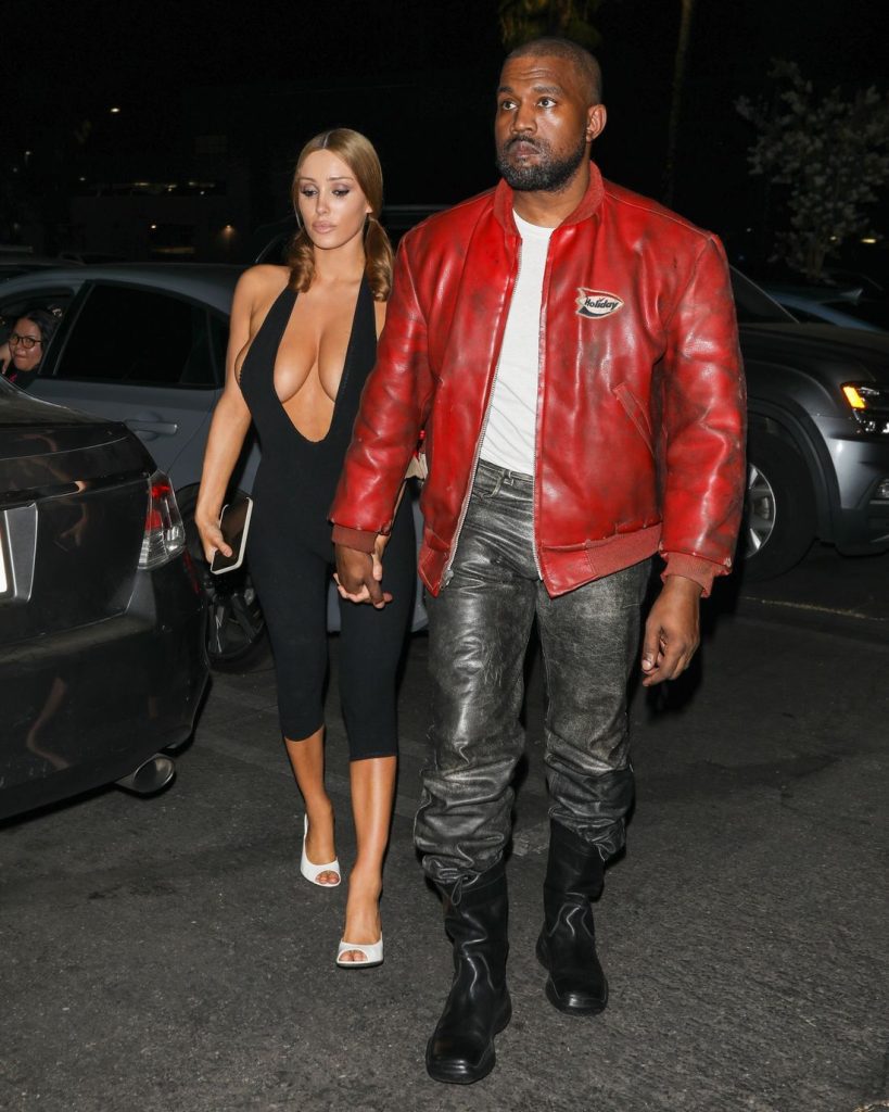 A woman in a black jumpsuit and white heels walks holding hands with a man wearing a red leather jacket, gray pants, and black boots at night in a parking lot #2.