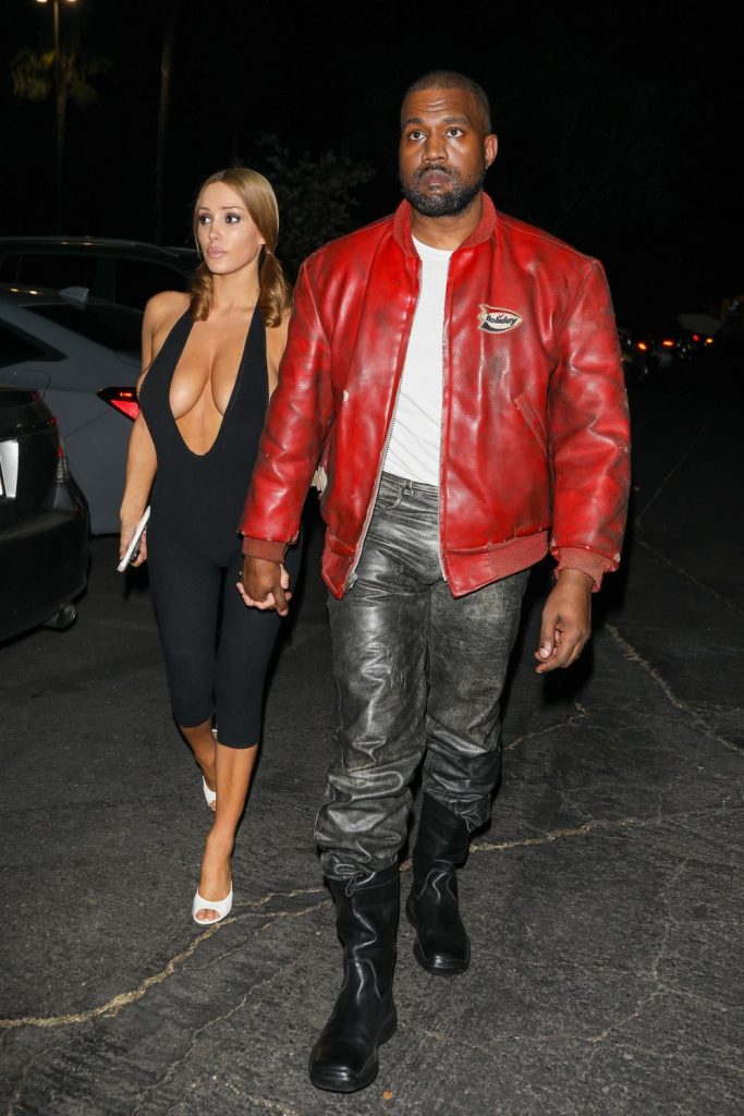 A woman in a black jumpsuit and white heels walks holding hands with a man wearing a red leather jacket, gray pants, and black boots at night in a parking lot #3.