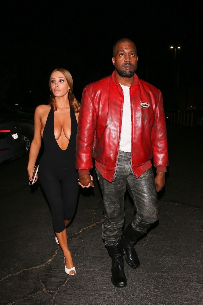 A woman in a black jumpsuit and white heels walks holding hands with a man wearing a red leather jacket, gray pants, and black boots at night in a parking lot #4.