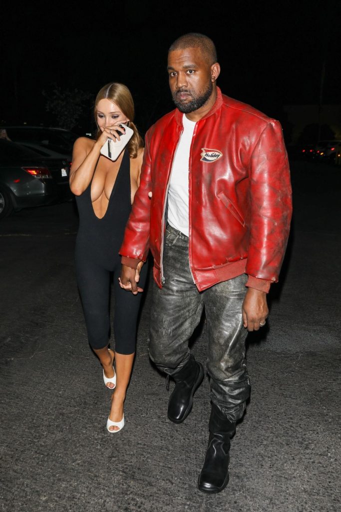 A woman in a black jumpsuit and white heels walks holding hands with a man wearing a red leather jacket, gray pants, and black boots at night in a parking lot #6.