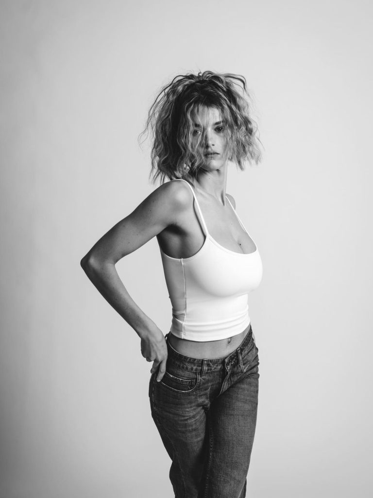 A black and white shot of a woman with messy blonde hair wearing a white tank top and blue jeans, posing with her hands on her hips.