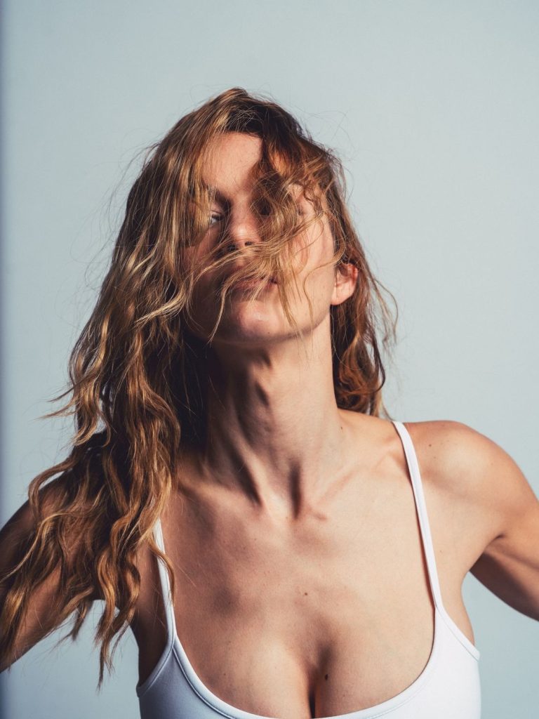 A woman with wavy, light brown hair wears a white tank top and poses against a plain background. Her hair partially covers her face, and she tilts her head slightly upward.