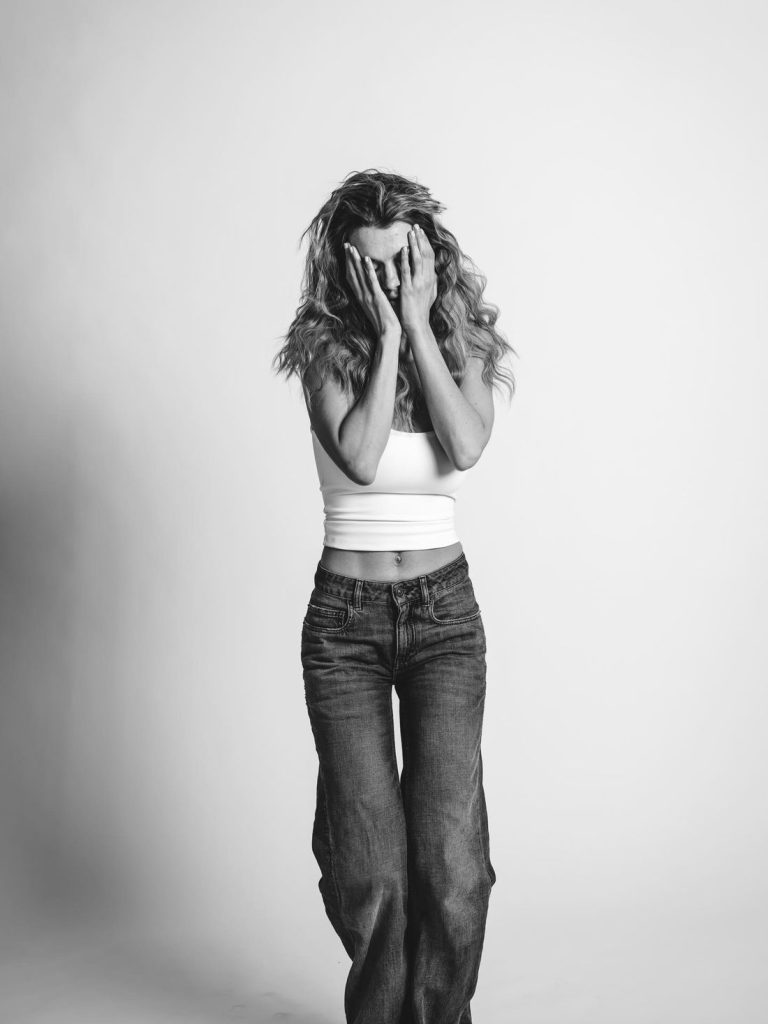 A woman wearing a white crop top and jeans stands against a plain background, covering her face with both hands. The image is in black and white.
