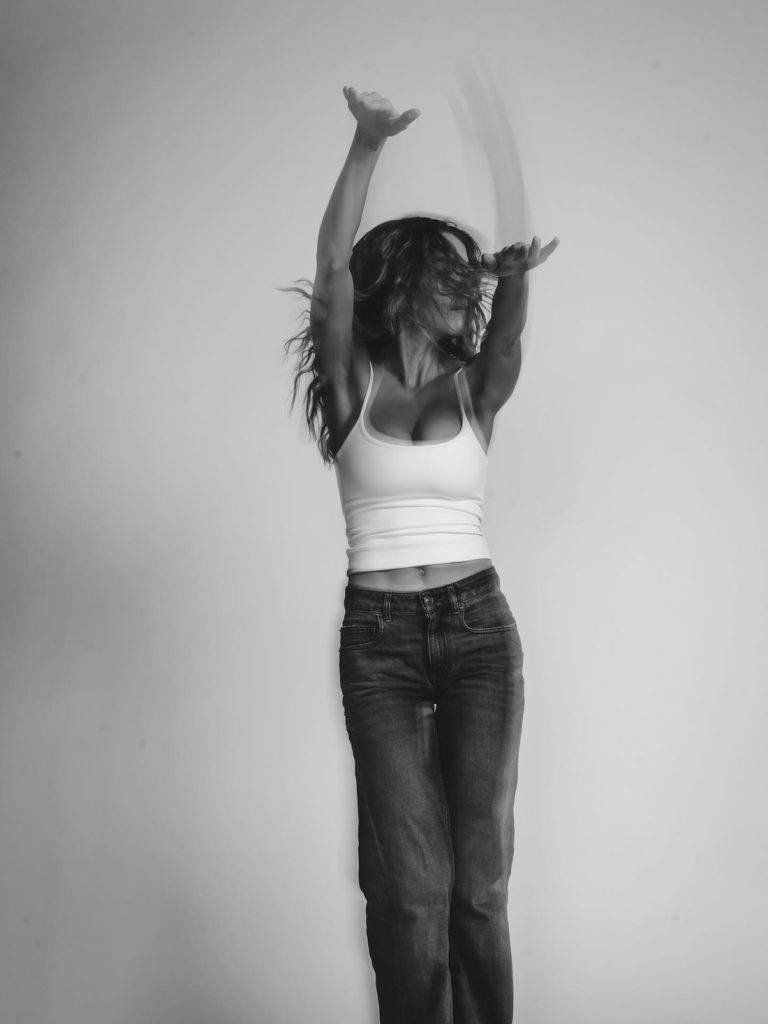 A woman in a white tank top and jeans dances energetically, her hair and arms in motion, against a plain light background in a black and white photo.