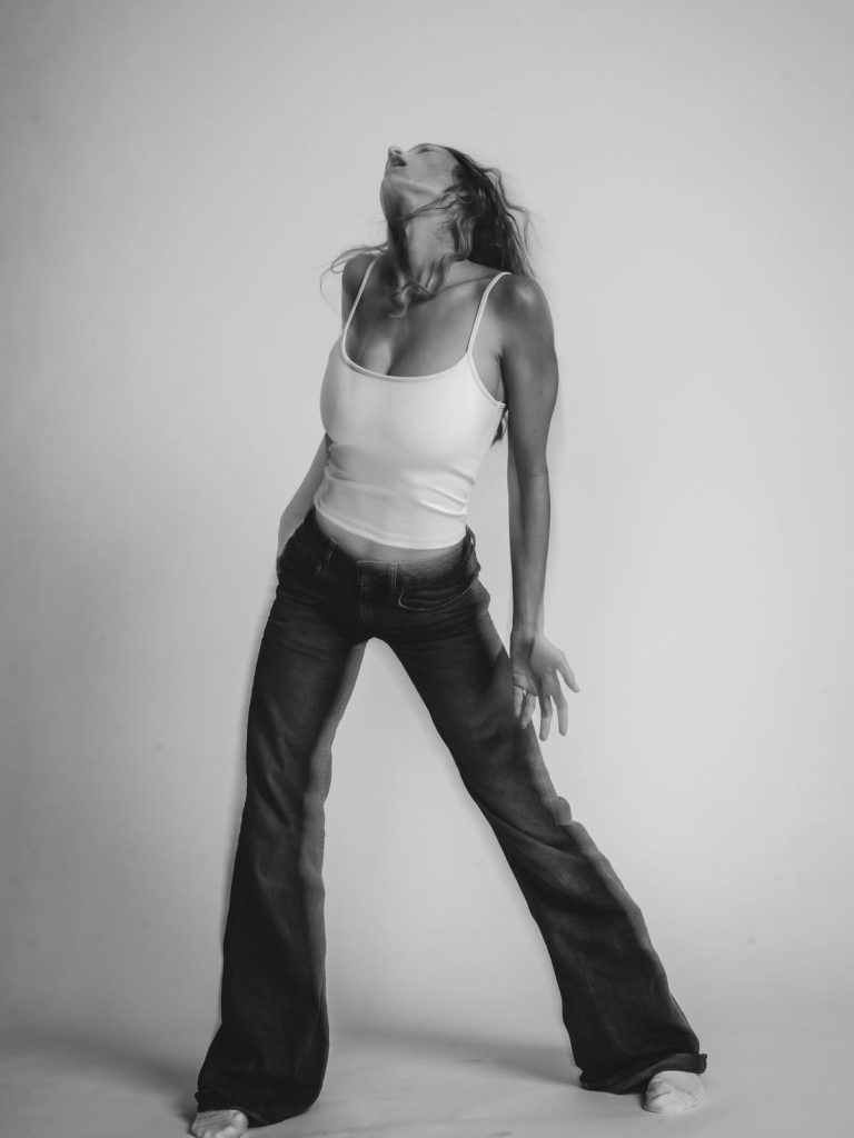 A woman wearing a white tank top and dark jeans stands with legs apart, head tilted back, and one hand on her hip in a dramatic pose against a plain, light background. The image is in black and white.