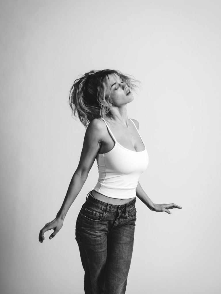 A woman wearing a white tank top and jeans stands with her eyes closed and head tilted back, as if dancing or moving fluidly, against a plain light background. The image is in black and white.