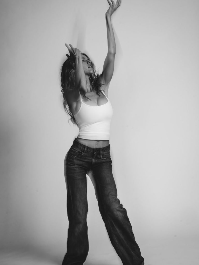 A woman in a white tank top and jeans poses gracefully with arms raised in a dance-like motion against a plain background. The image is in black and white, creating an artistic and expressive mood.