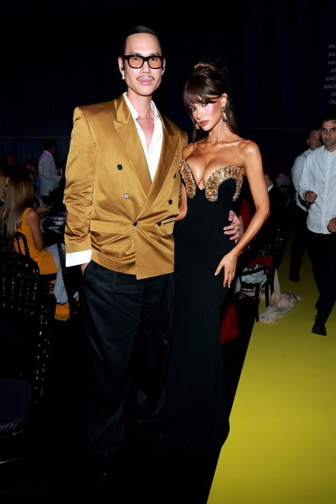 A man in a gold double-breasted blazer and glasses stands next to a woman in a black gown with a gold embellished bodice. They are posing together on a yellow carpet at a formal event with people in the background.