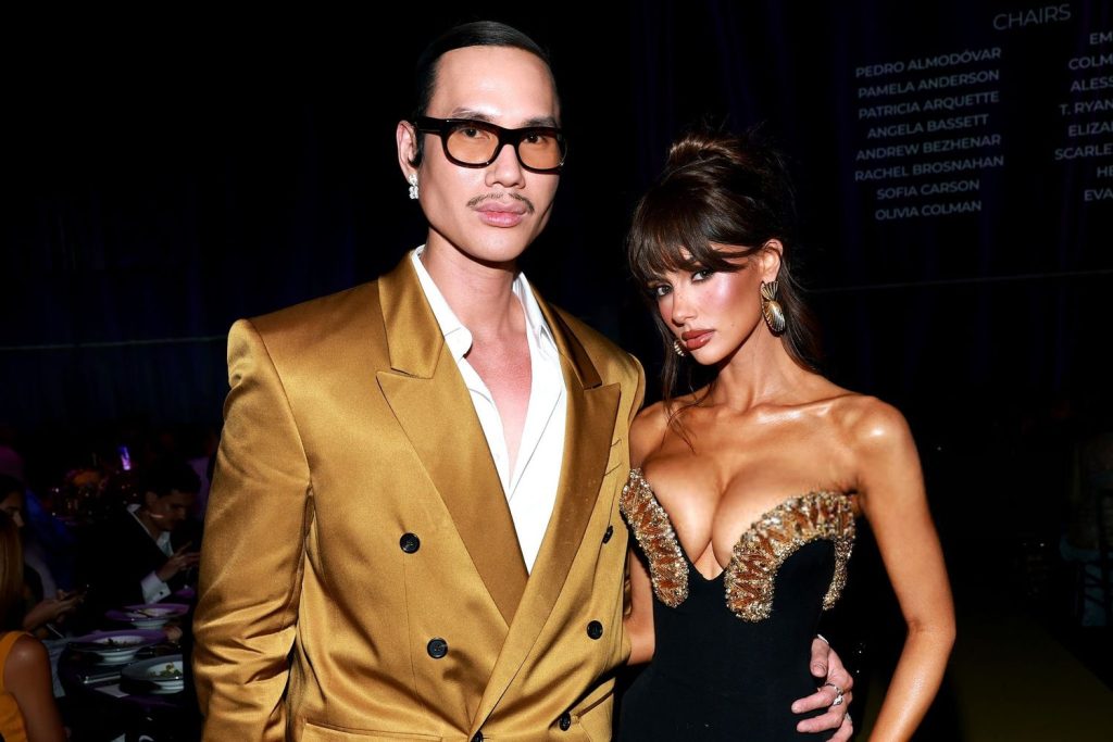 Close up of a man in a gold double-breasted blazer and glasses stands next to a woman in a black gown with a gold embellished bodice. They are posing together on a yellow carpet at a formal event with people in the background.
