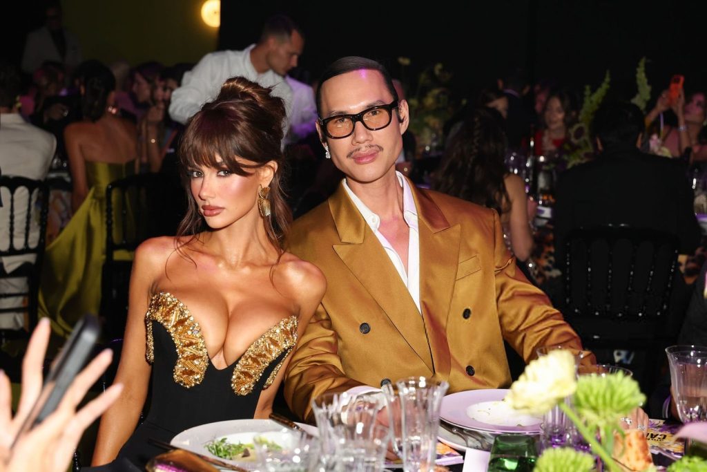 A woman in a black and gold dress and a man in a gold suit jacket and glasses sit together at a formal event, surrounded by elegantly dressed guests, tables with plates, glasses, and floral centerpieces.