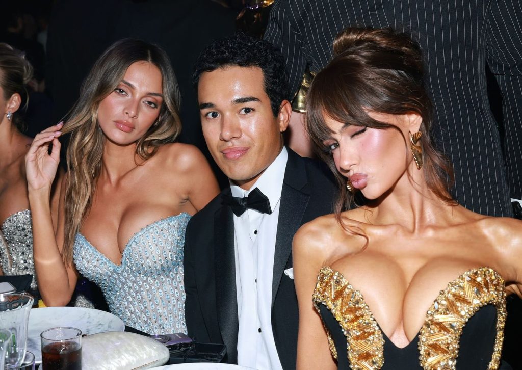 Three people dressed formally are seated at a table. Two women in sequined dresses flank a man in a black tuxedo. All are posing and looking at the camera amid a glamorous event setting.