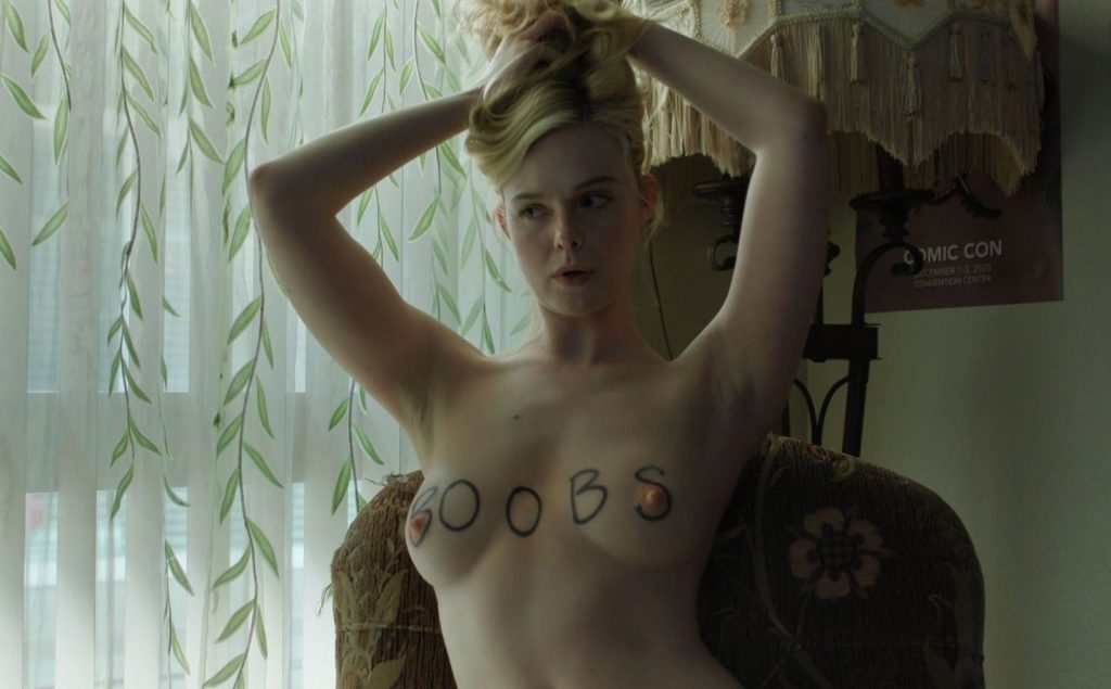 Elle Fanning topless with BOOBS written on her breasts. She twists and holds her hair up with both hands.