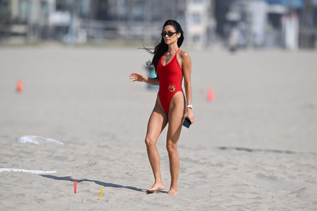 A brunette woman wearing a red swimsuit and sunglasses walks barefoot on a sandy beach, holding a phone in one hand. The background is blurred with scattered cones and beach structures visible.