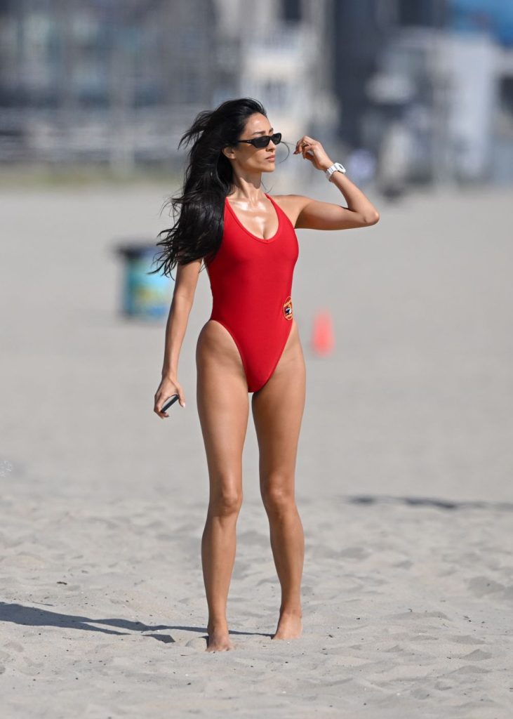 A woman wearing a red one-piece swimsuit and sunglasses stands barefoot on a sandy beach, holding a phone, with a blurred background of beach objects and buildings.
