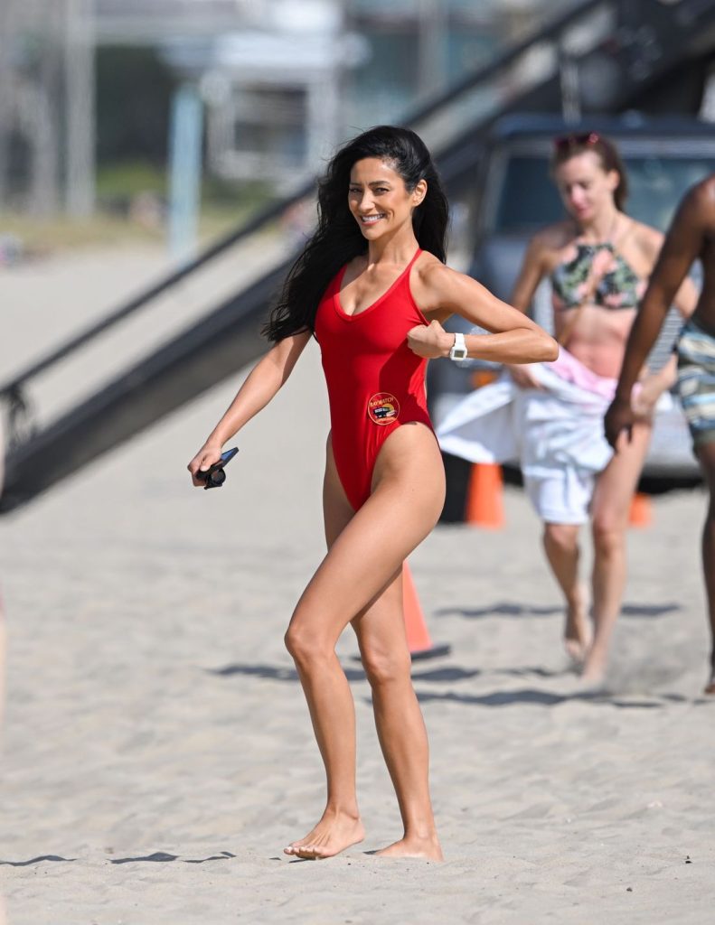 A woman in a red one-piece swimsuit walks barefoot on a sandy beach, smiling and holding something in her right hand. Other people in swimwear are visible in the background.