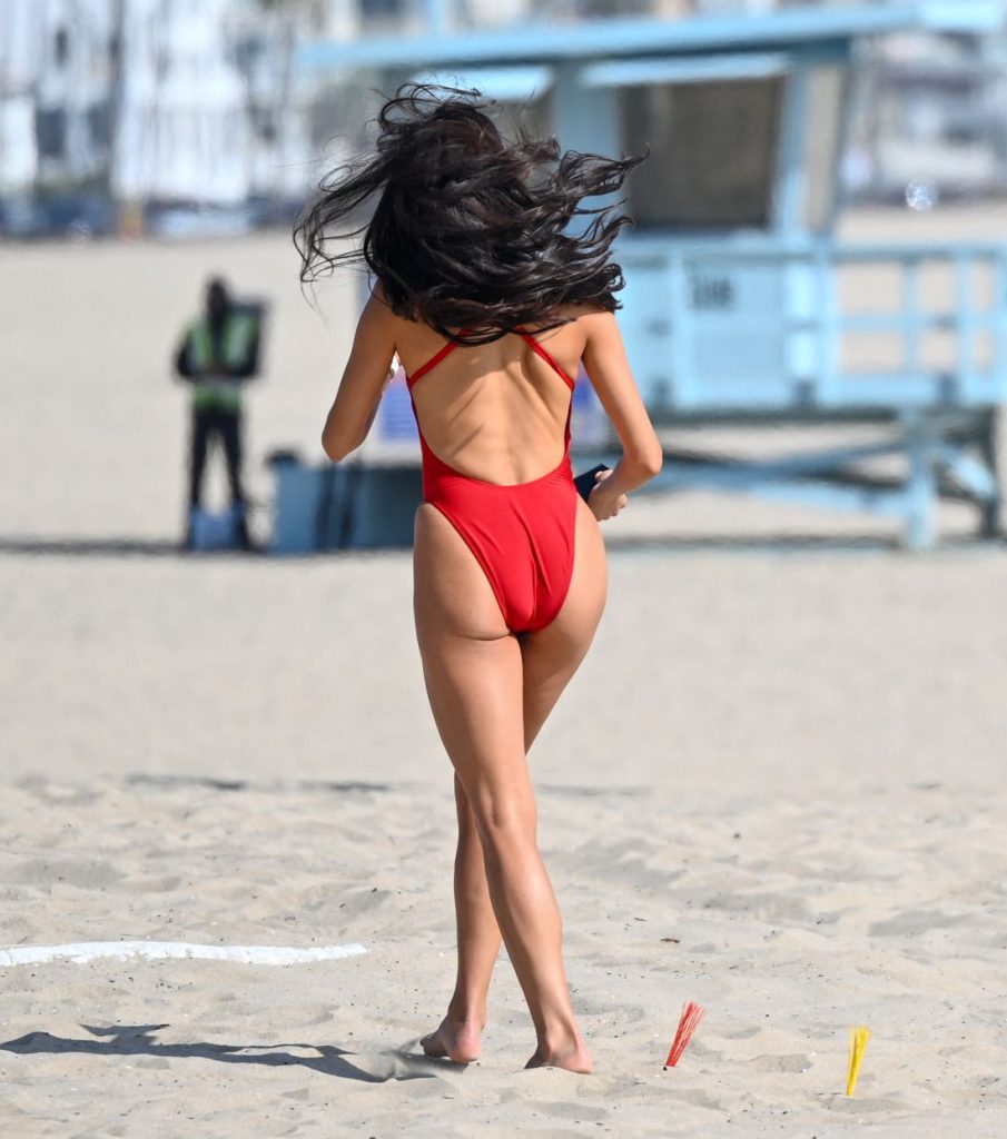 Rear view of a woman with long dark hair, wearing a red one-piece swimsuit, walks on a sandy beach toward a blue lifeguard tower. The scene is sunny and appears relaxed, with another person blurred in the background.