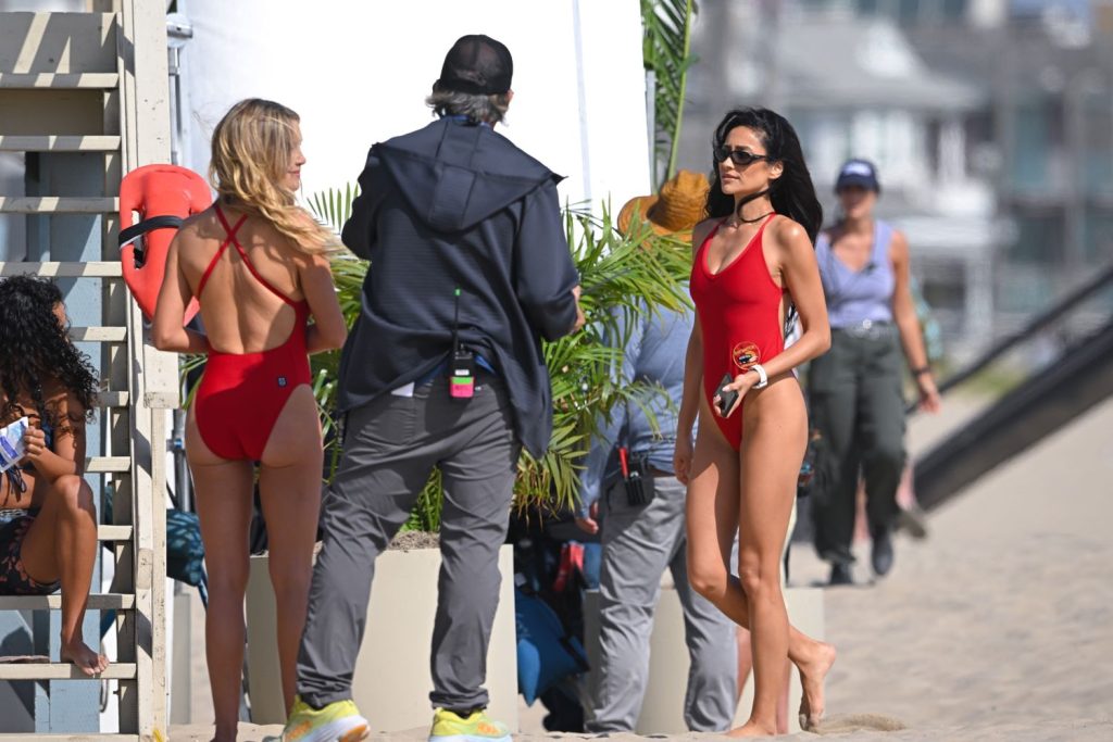 Two women in red swimsuits stand on a sandy beach near a man wearing a jacket and cap, with other people and buildings visible in the background. One woman holds sunglasses, and there are palm plants nearby.