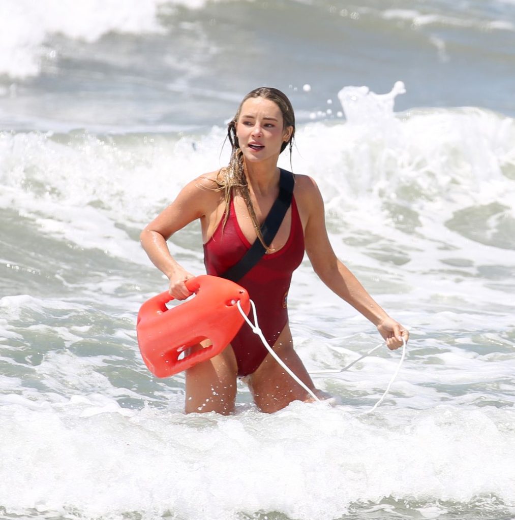 A woman in a red swimsuit stands in the ocean waves, holding a red lifeguard rescue buoy and wearing a black strap across her torso. Water splashes around her as she looks ahead.