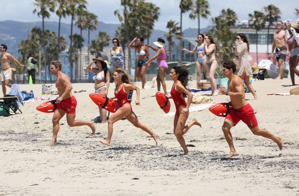 Four lifeguards in red swimsuits run toward the ocean on a sandy beach, holding rescue cans, while a crowd of beachgoers watches in the background under a sunny sky with palm trees #3.