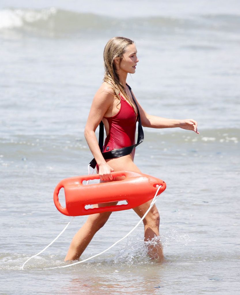 A woman in a red swimsuit walks through shallow ocean water, carrying an orange lifeguard rescue float and wearing a black strap over her shoulder.