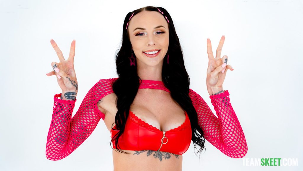 A woman with long dark hair, wearing a red fishnet crop top and matching red top, smiles while holding up both hands in a peace sign gesture; the background is plain white.