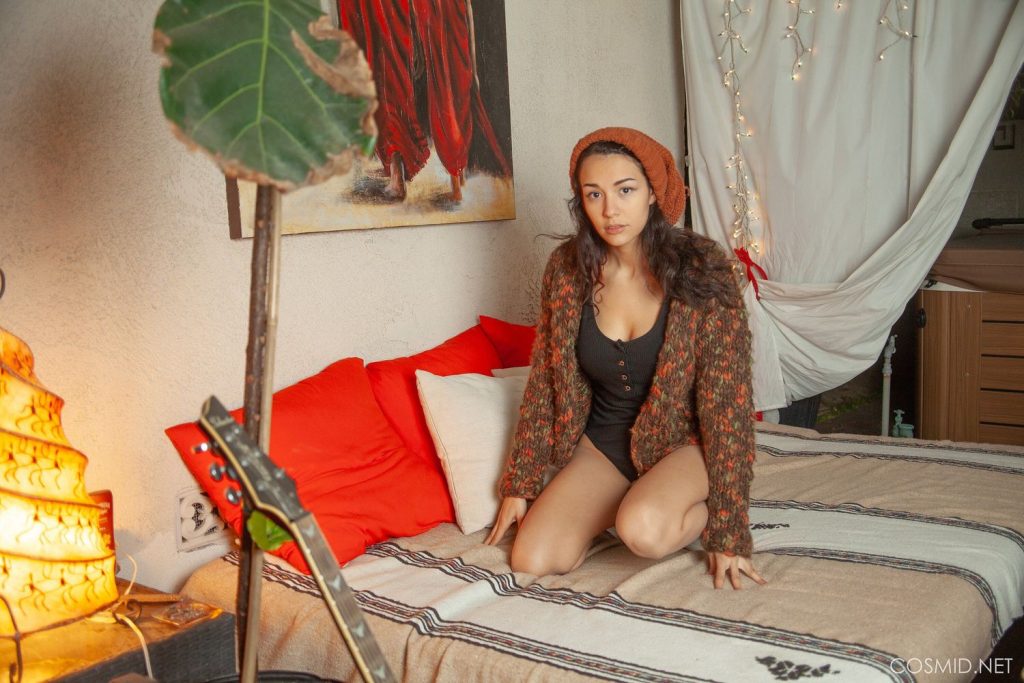 A woman wearing a brown knit hat and cardigan sits on a bed with red pillows in a cozy, warmly lit room decorated with string lights, a large leaf, and a painting in the background.