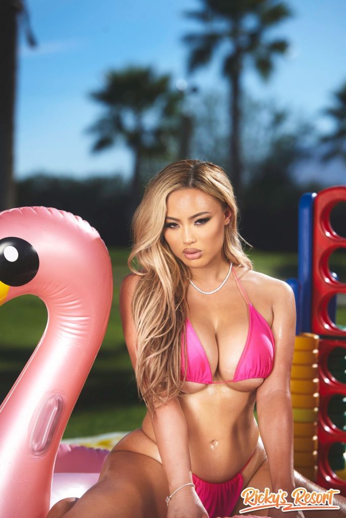 Asian woman in a pink bikini sits next to a large inflatable flamingo in an outdoor setting with palm trees and lawn games in the background. The image is bright and summery.