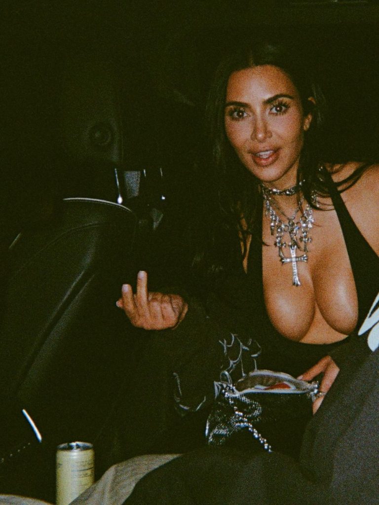 A woman with long dark hair, wearing a black outfit and layered silver necklaces, sits in a dimly lit car and holds up her middle finger. A can is visible at her side.