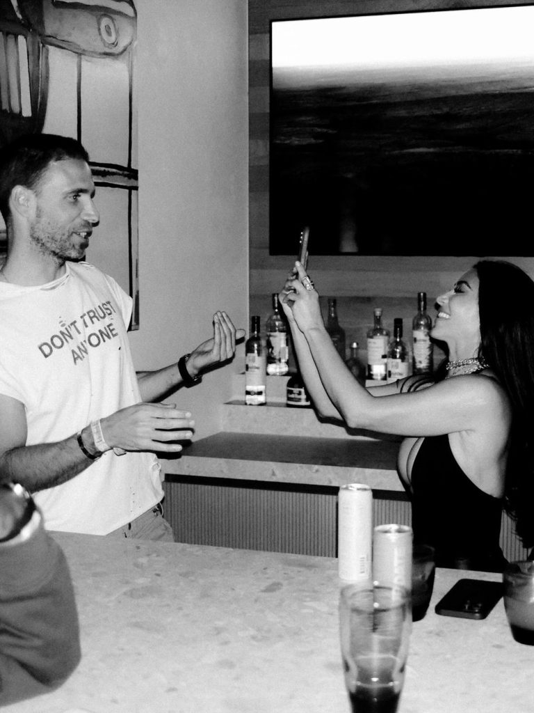 A man and a woman interact at a bar; the man gestures while the woman, smiling, takes his photo with her phone. Various bottles and drinks are visible in the background. The scene is in black and white.