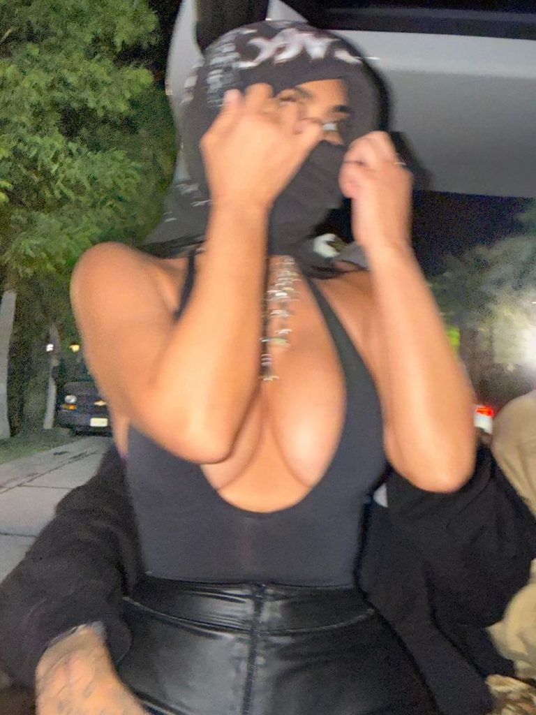 A woman wearing a sleeveless black top and leather pants adjusts a hood or scarf over their head and face while sitting in a car at night. The image is slightly blurred, suggesting motion.
