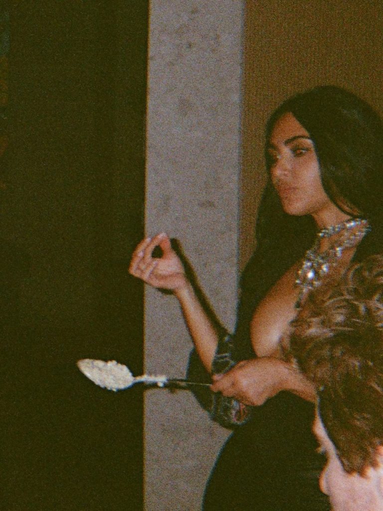 A woman with long dark hair, wearing a low-cut dress and a statement necklace, holds a serving spoon with food in one hand at what appears to be an indoor gathering.
