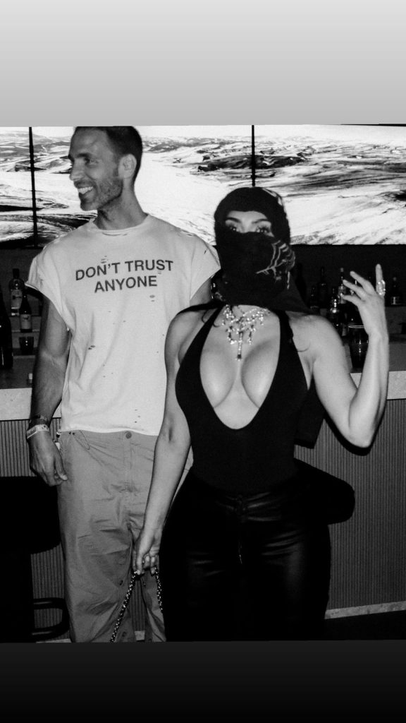 A black and white photo of a smiling man in a DONT TRUST ANYONE T-shirt standing next to a woman with a headscarf covering her face, wearing a low-cut top and jewelry, raising her hand in a gesture. They are in front of a bar.