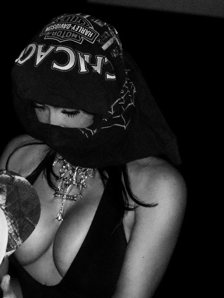 A woman in a black top with a low neckline wears a Harley-Davidson bandana over their head and face, adorned with layered chain necklaces, including a large cross. The image is in black and white.