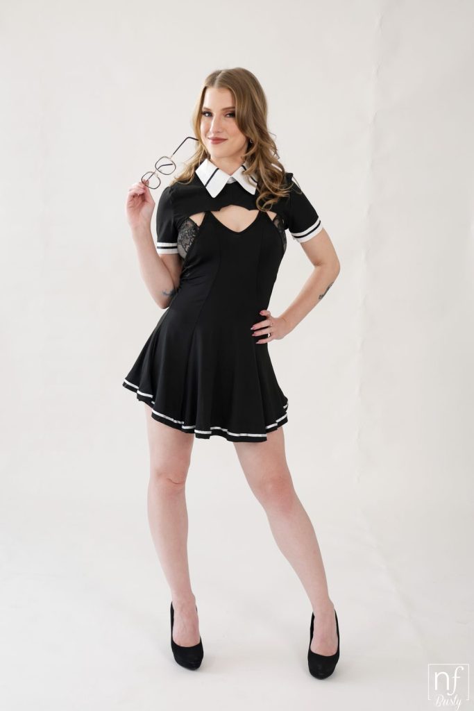 A woman in a black dress with white trim and a white collar poses confidently, holding eyeglasses in one hand. She stands on a white backdrop, wearing black high heels and smiling at the camera.