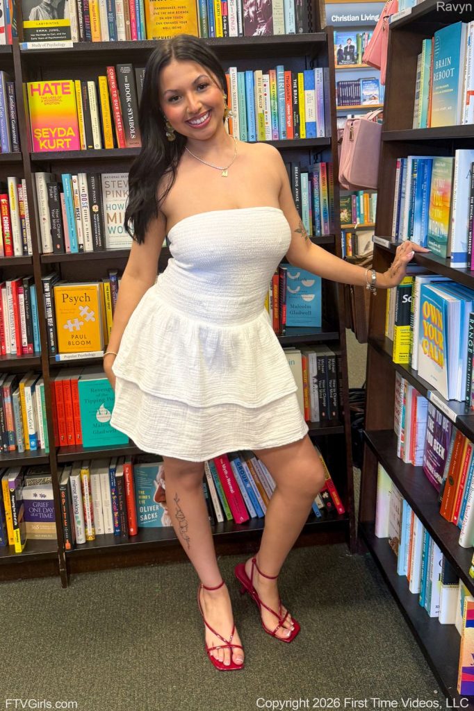 A woman in a white strapless dress and red shoes poses smiling between bookshelves in a bookstore. The shelves are filled with colorful books, and she stands with one hand resting on the shelf.